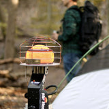 Portable heater with cage on a stand in a forest setting, person with backpack in background