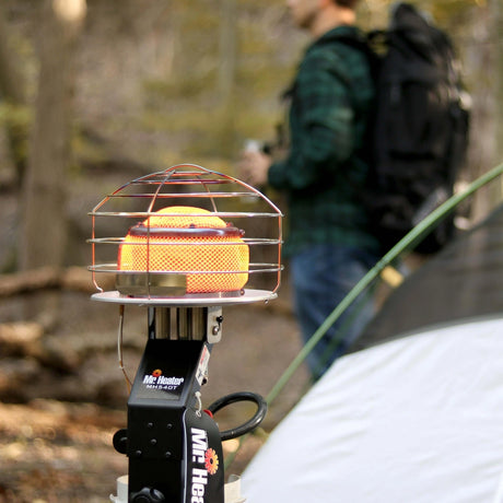Portable heater with cage on a stand in a forest setting, person with backpack in background
